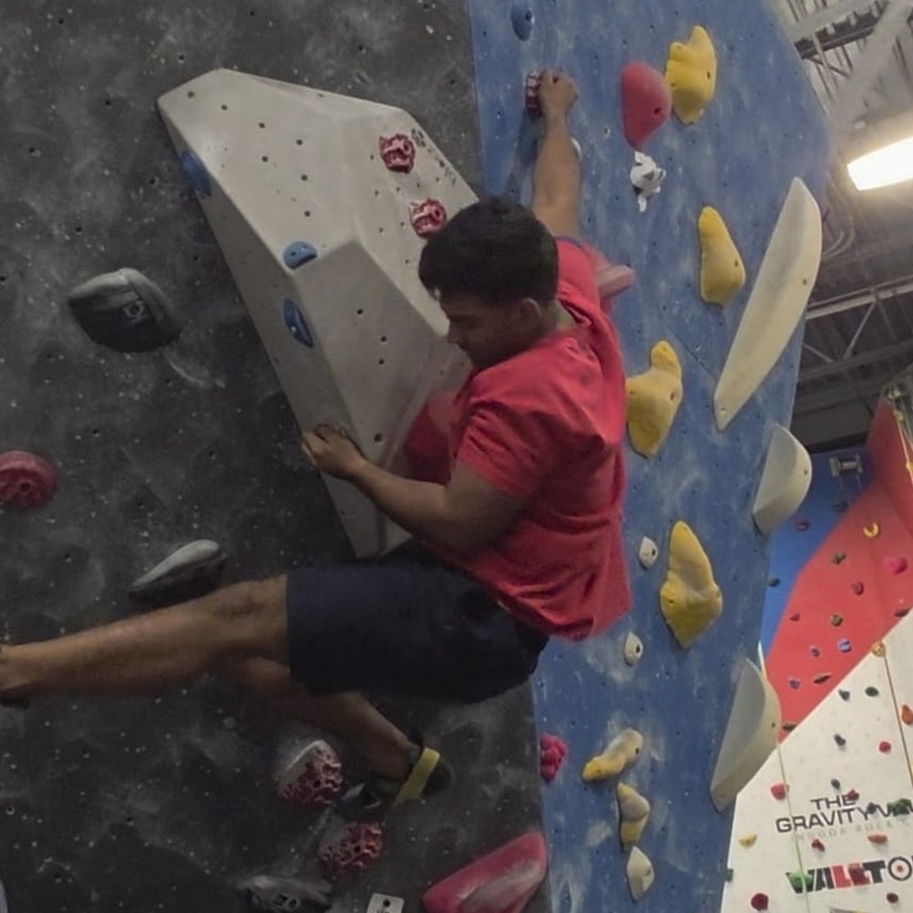 Ashish mid-move on a steep overhang at an indoor bouldering gym, body horizontal, reaching for the next hold